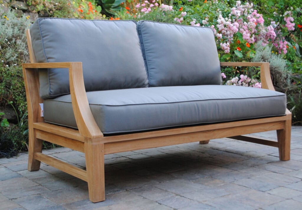 side angle view of teak Capitola Deep Seating Loveseat with gray Cushions with flowers in background Paradise Teak Bay Area California