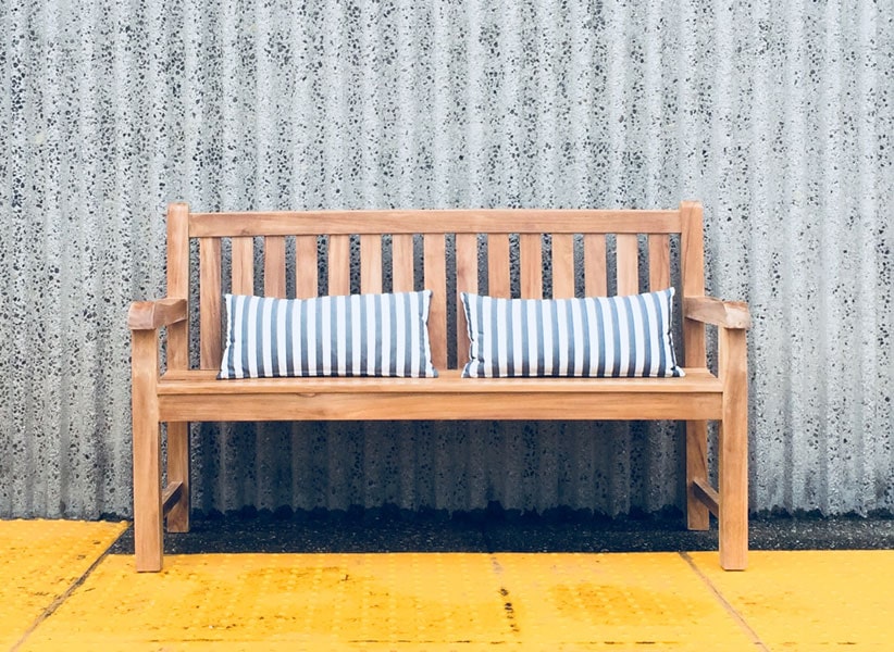 teak outdoor bench with two blue and white accent pillows sits against a concrete wall Paradise Teak Bay Area California